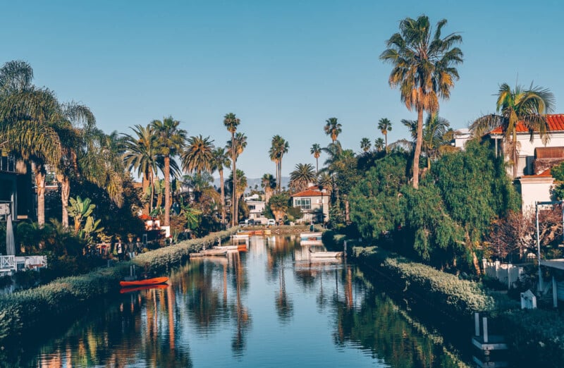 Un canal pittoresque bordé de palmiers luxuriants et d’une végétation vibrante se reflète sur une eau calme. De charmantes maisons bordent le canal, avec de petits bateaux amarrés le long des côtés. Le ciel est clair et bleu, créant une atmosphère sereine et tropicale.