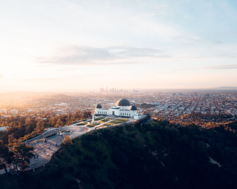 Vue aérienne de l'observatoire Griffith perché sur une colline, avec le paysage urbain tentaculaire de Los Angeles en arrière-plan sous un ciel de lever de soleil doux et lumineux. Les dômes emblématiques de l'observatoire se détachent sur le paysage urbain.