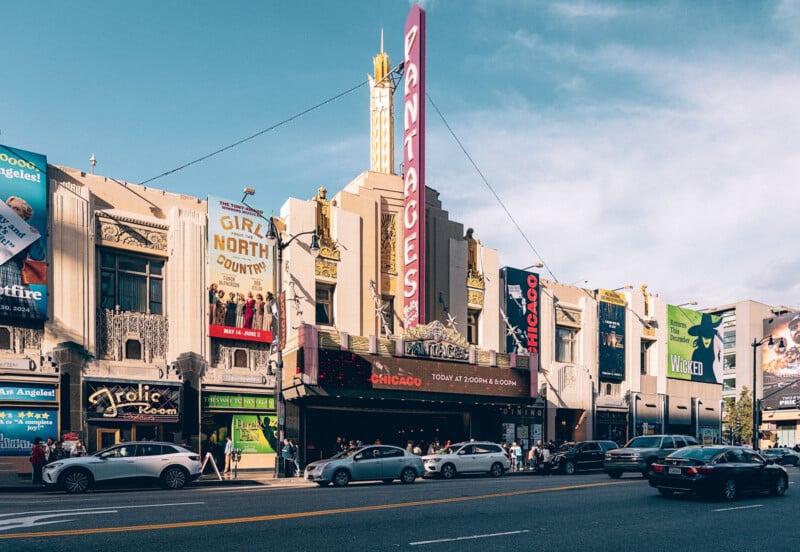 Une rue animée de la ville avec le Théâtre Pantages, qui met en évidence le chapiteau de "Chicago." Les bâtiments environnants arborent de grandes banderoles colorées pour divers spectacles. Les voitures et les piétons sont visibles dans la rue.