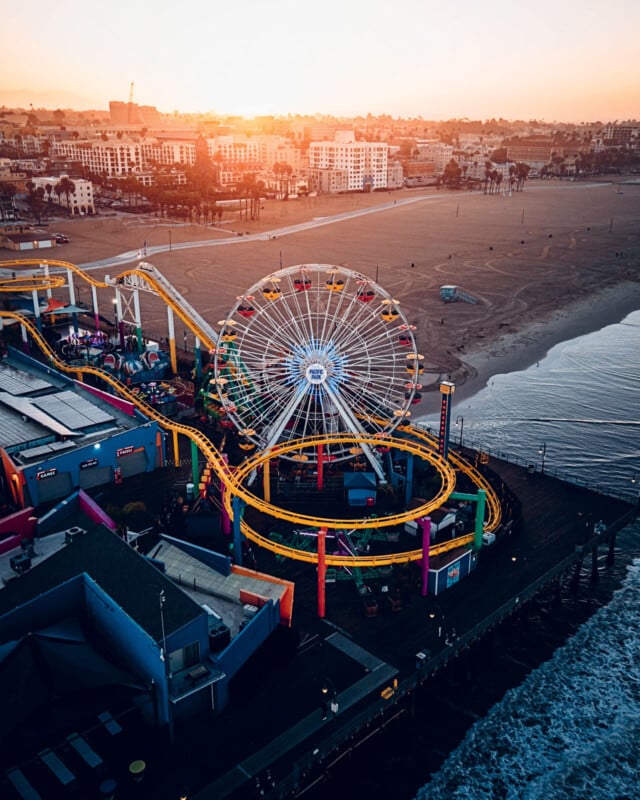 Vue aérienne d'un parc d'attractions sur une jetée en bord de mer au coucher du soleil. Une grande roue et des montagnes russes sont mises en évidence. La plage s'étend au loin, avec un paysage urbain visible en arrière-plan.