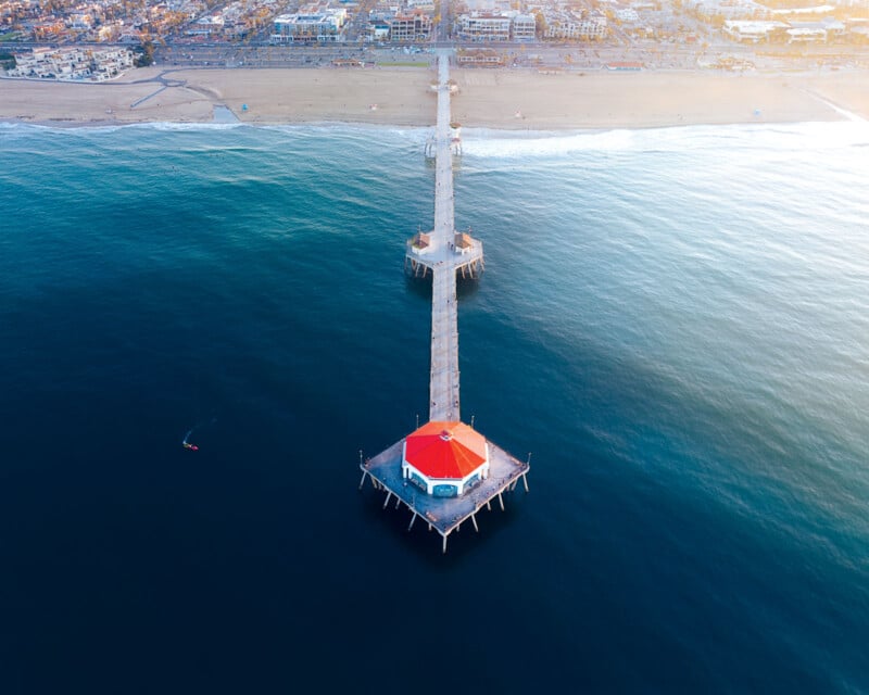 Vue aérienne d'une longue jetée s'étendant dans l'océan, avec à son extrémité un bâtiment au toit rouge. Le rivage est bordé de sable et de bâtiments, et une personne seule pagaye dans l’eau près de la jetée.