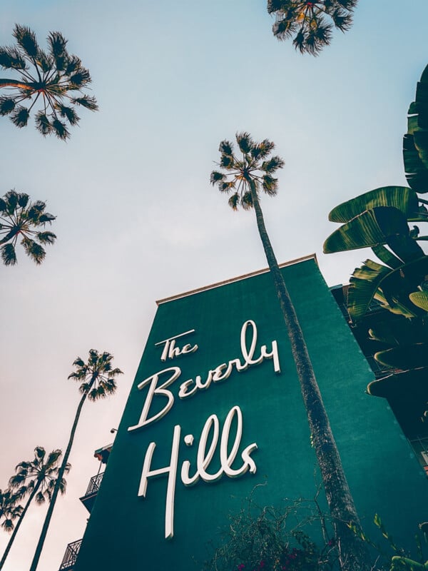 De grands palmiers entourent un bâtiment vert avec une pancarte rétro indiquant "Les Beverly Hills." Le ciel est clair, ajoutant à l’ambiance californienne classique.