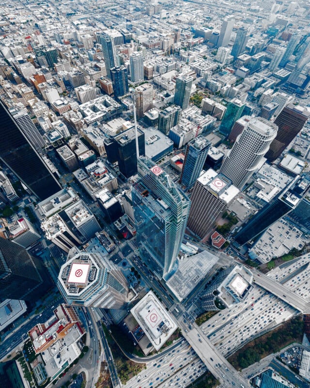 Vue aérienne d'une ville animée avec de grands gratte-ciel, dont une imposante tour de verre. Des routes à fort trafic entourent les bâtiments et plusieurs héliports sur les toits sont visibles. Le paysage urbain s'étend au loin.