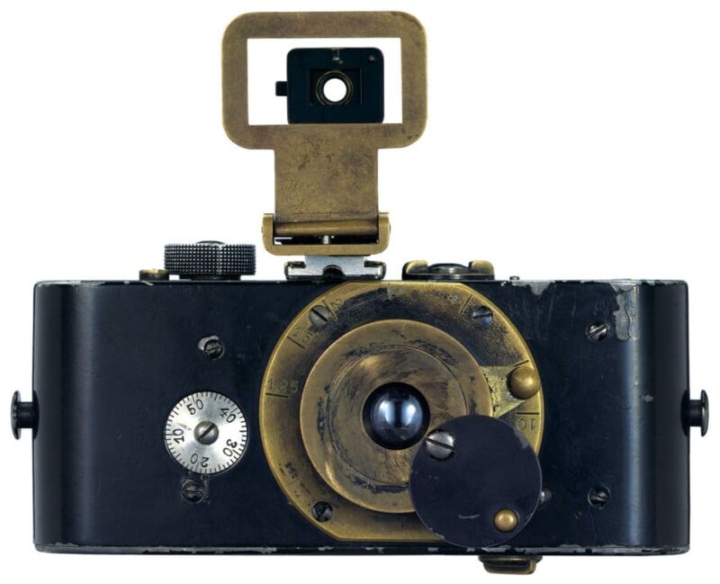 A vintage camera with a brass lens and a folding viewfinder. The camera has a weathered, metal body with a small dial on the left. The lens is prominently centered, showcasing an intricate, aged design.