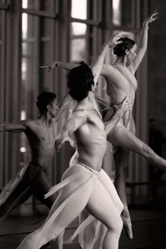 A black and white photo of three ballet dancers in motion. Two men and one woman, all in elegant poses, wearing form-fitting attire. They are captured mid-performance with a background of tall windows.