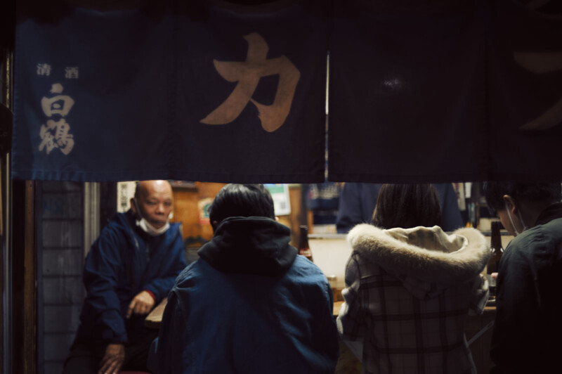 A small, dimly lit Japanese bar with people sitting close together on stools. A server stands in the background. Dark blue curtains with Japanese characters hang above, creating a cozy, intimate ambiance.