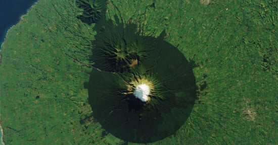 Aerial view of a snow-capped volcano surrounded by lush green vegetation. The dark, radial pattern of forest growth contrasts with the vibrant greenery. The coastline is visible to the left of the image.