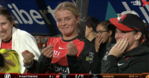 Cricket players in team uniforms are seen on a bench, making playful gestures towards the camera. The score is shown partially at the bottom of the screen.