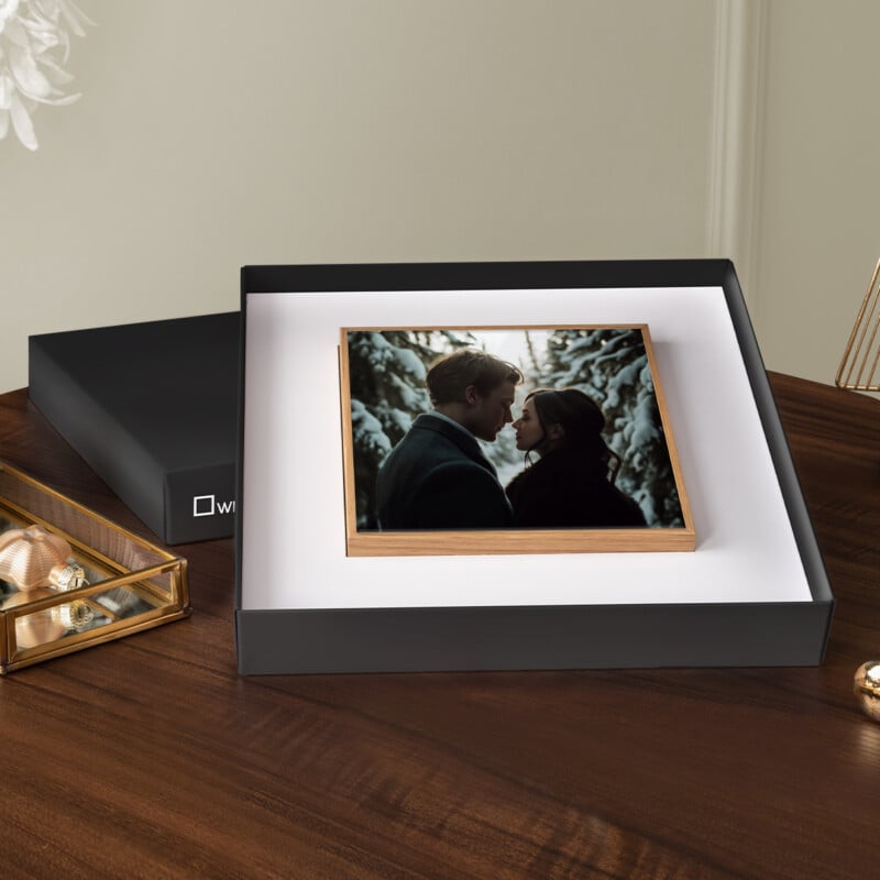 A sleek black gift box lies open on a wooden table, revealing a framed photo of a couple sharing an intimate moment outdoors. The scene is set against a wintry backdrop. Nearby, festive decorations add a touch of elegance.