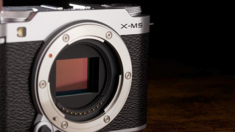 A close-up view of a silver and black digital camera with the lens removed, revealing the sensor. The camera has textured grips and the label "X-M5" near the top. The background is dark.