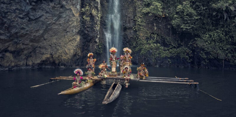 A group of people wearing traditional, colorful costumes and headdresses stand and sit on a raft before a waterfall, surrounded by lush greenery and water. The scene conveys a sense of cultural celebration and natural beauty.