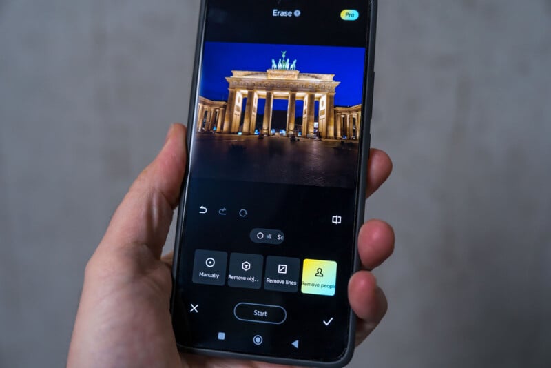 A person holding a smartphone displaying an image editing app. The screen shows a night photo of an illuminated landmark with columns and statues, possibly in a historic architectural style. Various editing tools are visible below the image.