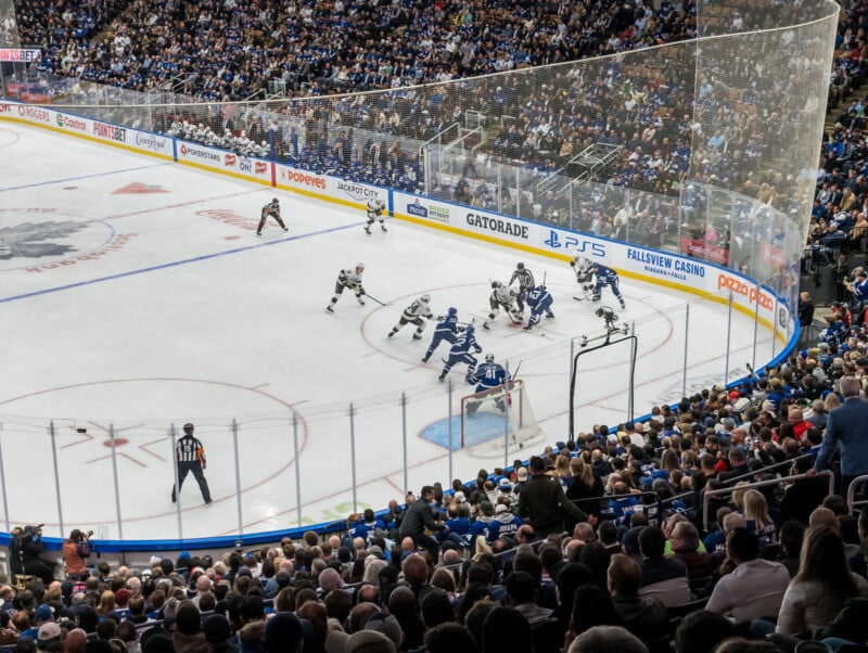 A lively hockey game in a packed arena. Players from two teams, wearing white and blue jerseys, are actively competing for the puck near one of the goals. Fans fill the stands, watching the action intently. An official observes the play closely.