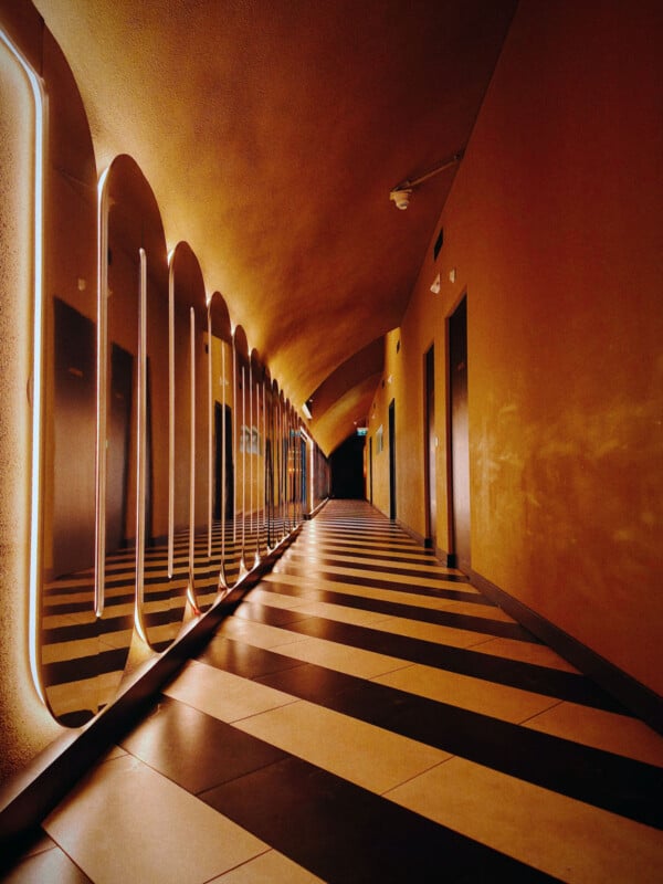 A long, narrow corridor with a checkered black and white floor and illuminated mirrors on the left wall. The right wall is plain, and the ceiling arches slightly. The lighting creates a warm, modern ambiance.