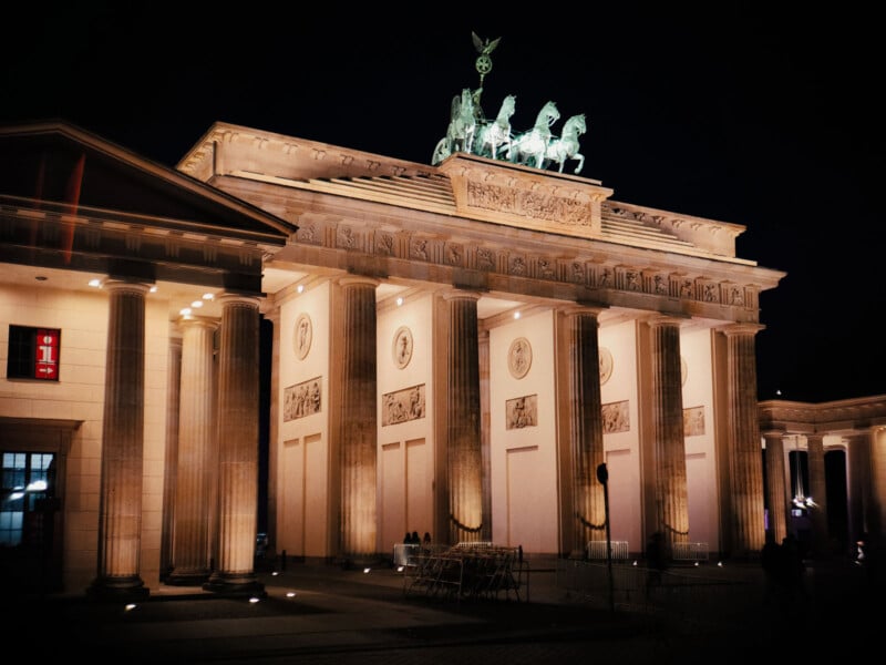 The image shows the Brandenburg Gate illuminated at night. The neoclassical monument features columns and a chariot statue on top, set against a dark sky.