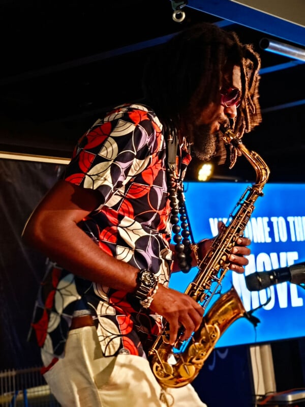 A musician with dreadlocks plays the saxophone passionately on stage. He's wearing sunglasses and a colorful patterned shirt. A bright sign in the background partially reads "Welcome to the Move.