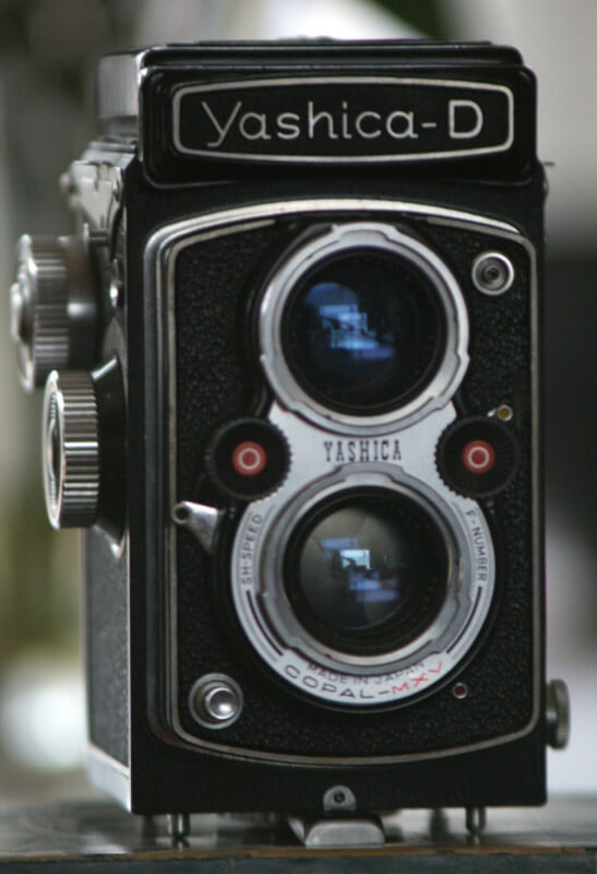 Close-up of a vintage Yashica-D twin-lens reflex camera, featuring two stacked lenses and various dials on a black body. The camera has silver accents and a textured grip, evoking a classic, retro aesthetic.