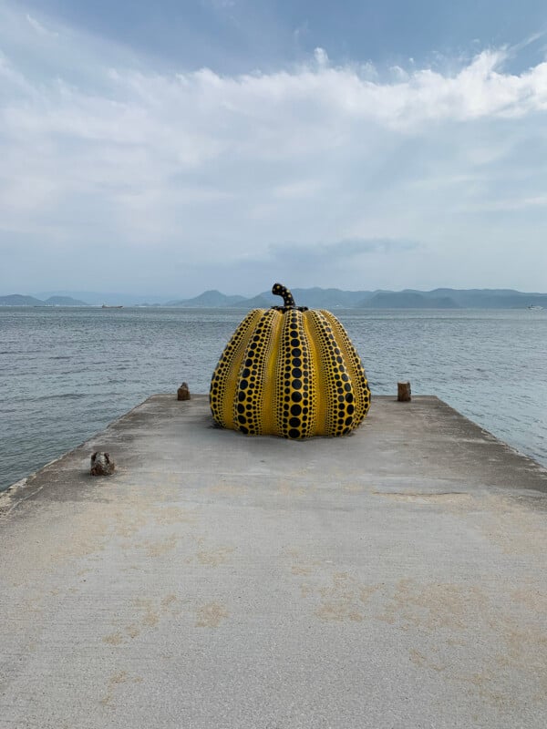 Une grande sculpture de citrouille jaune à pois noirs se trouve sur une jetée en béton, surplombant une mer calme. L’arrière-plan présente des montagnes lointaines sous un ciel partiellement nuageux.