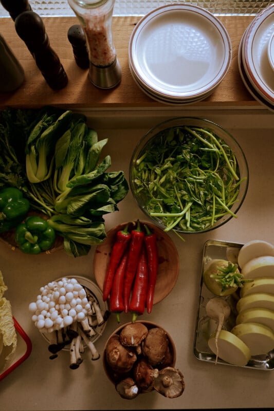 A kitchen counter displays a variety of fresh vegetables, including green peppers, bok choy, a bowl of leafy greens, red chili peppers, mushrooms, and sliced kohlrabi. Plates are stacked nearby, ready for use.