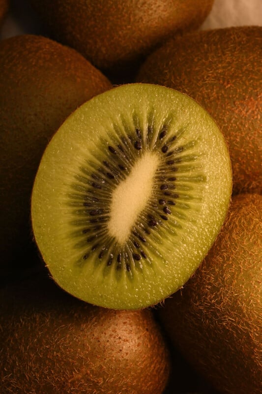 Close-up of a halved kiwi fruit displaying its vibrant green flesh and tiny black seeds, set against a background of whole kiwi fruits with fuzzy brown skin.