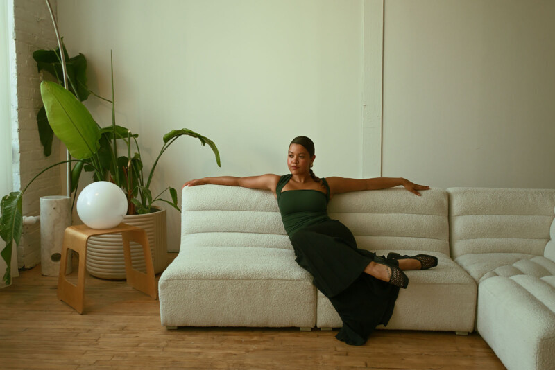 A person wearing a green top and black pants is lounging on a white sofa in a modern room with wooden flooring. Large green plants and a round white lamp on a wooden stool are arranged in the background.