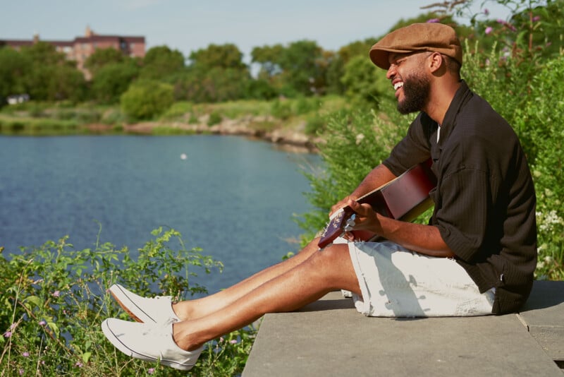 A man sits by a lake, wearing a brown cap and holding a guitar. He is smiling while seated on a stone ledge, surrounded by green foliage. His legs are stretched out, wearing white sneakers and light shorts on a sunny day.