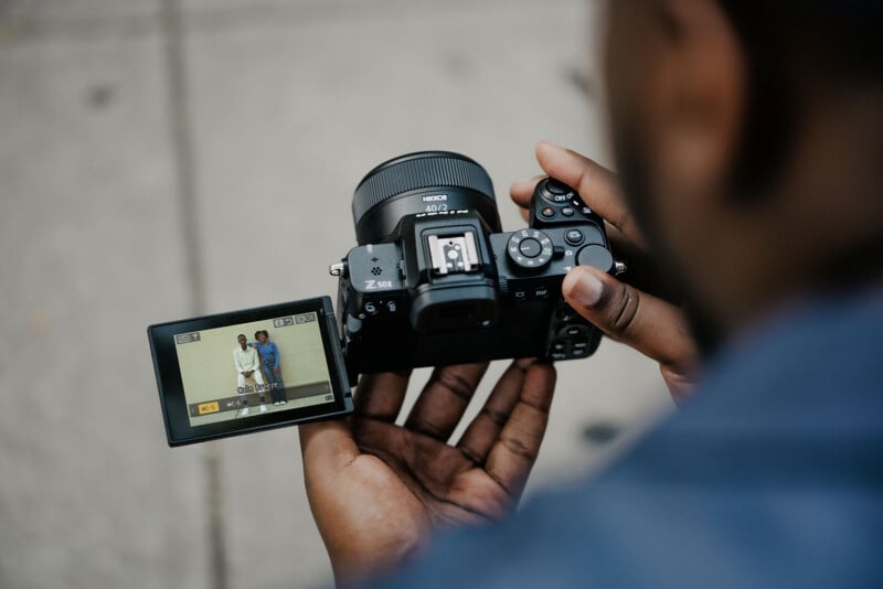 A person holds a camera with a flip-out screen showing a live view of two people standing side by side. The camera is held against a blurred outdoor background. The scene is captured from over the shoulder of the person holding the camera.