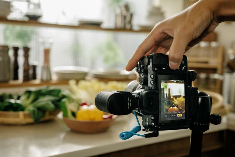 A hand holding a camera capturing a close-up of a bowl of colorful vegetables on a kitchen counter. Various veggies like peppers and greens are visible in the soft-focus background.