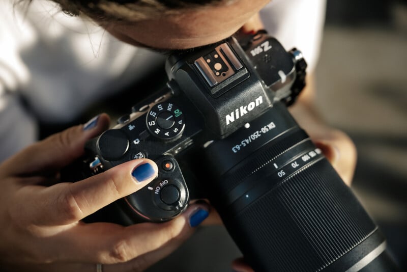 A person with blue nail polish holds a Nikon camera, looking through the viewfinder. The camera lens and dial settings are visible, capturing a moment of focused photography.