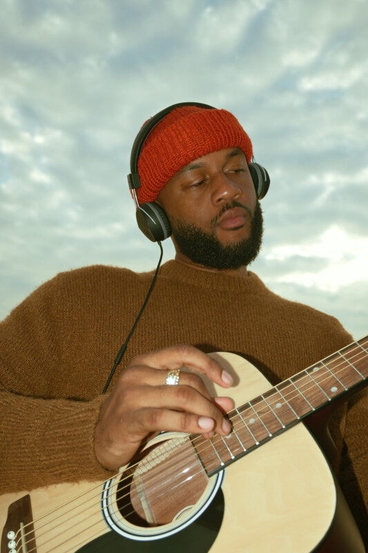 A man is playing an acoustic guitar while wearing headphones and a red beanie. He's dressed in a brown sweater, standing against a cloudy sky backdrop.