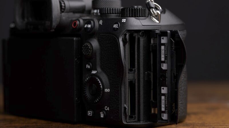 A close-up of a digital camera showing two empty memory card slots with detailed camera controls visible, including buttons for AEL, ISO, and others. The image highlights the camera's textured design and metal ring on a dark background.