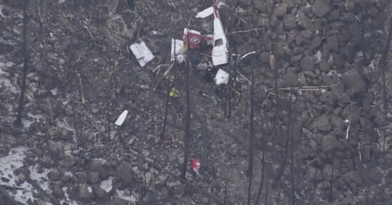 Aerial view of a plane crash site in a rocky, barren area. Debris from the aircraft is scattered among the rocks, with some parts still recognizable. The surrounding area appears charred, indicating possible fire damage.