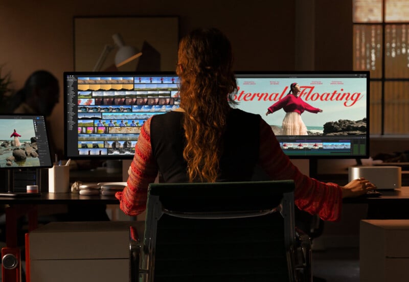 A person with long hair sits at a desk, editing a video on two large monitors. The screen shows video clips and a frame with the words "Floating Eternal" featuring a person in a red dress on rocks. The room is dimly lit, with various tech equipment around.