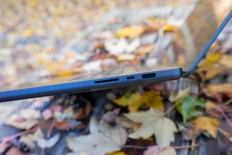 A laptop with a thin profile is partially open and placed on an outdoor surface covered with yellow and brown fallen leaves. The image showcases the laptop's ports and sleek design.