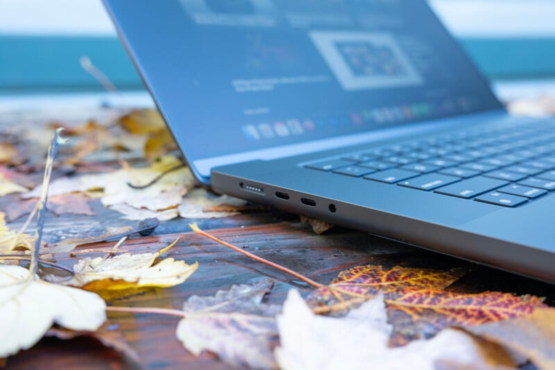 A laptop on an outdoor wooden surface covered with fallen autumn leaves. The screen displays a blurred image, and the keyboard is partially visible. The focus is on the laptop's side ports.