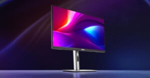 A sleek computer monitor with a vibrant, colorful display stands on a reflective surface. The screen shows a dynamic abstract background with shades of pink, purple, and blue. The monitor has a thin bezel and a sturdy stand.