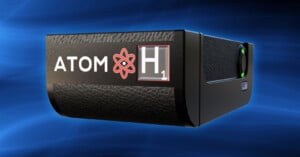 A black device labeled "ATOM" with a red atom symbol and "H1" text on a blue background. The device has a textured surface and a glowing green button.