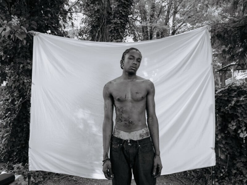 A black and white photo of a shirtless person standing in front of a white backdrop, surrounded by dense foliage. The person appears relaxed, with their hands in their pockets and their hair styled in cornrows.