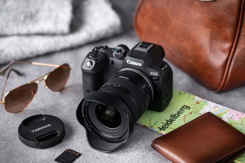 A Canon EOS R7 camera with a Tamron lens sits on a map labeled "Heidelberg." Nearby are a pair of sunglasses, a camera lens cap, an SD card, a brown leather wallet, and a brown leather bag. A gray knit fabric is in the background.