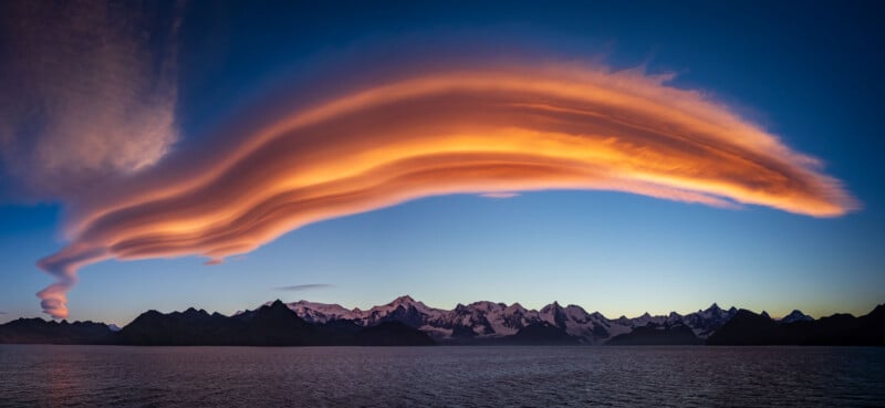 A striking lenticular cloud, glowing orange from the setting sun, stretches across a deep blue sky above a dark silhouette of a mountain range, with calm water in the foreground.