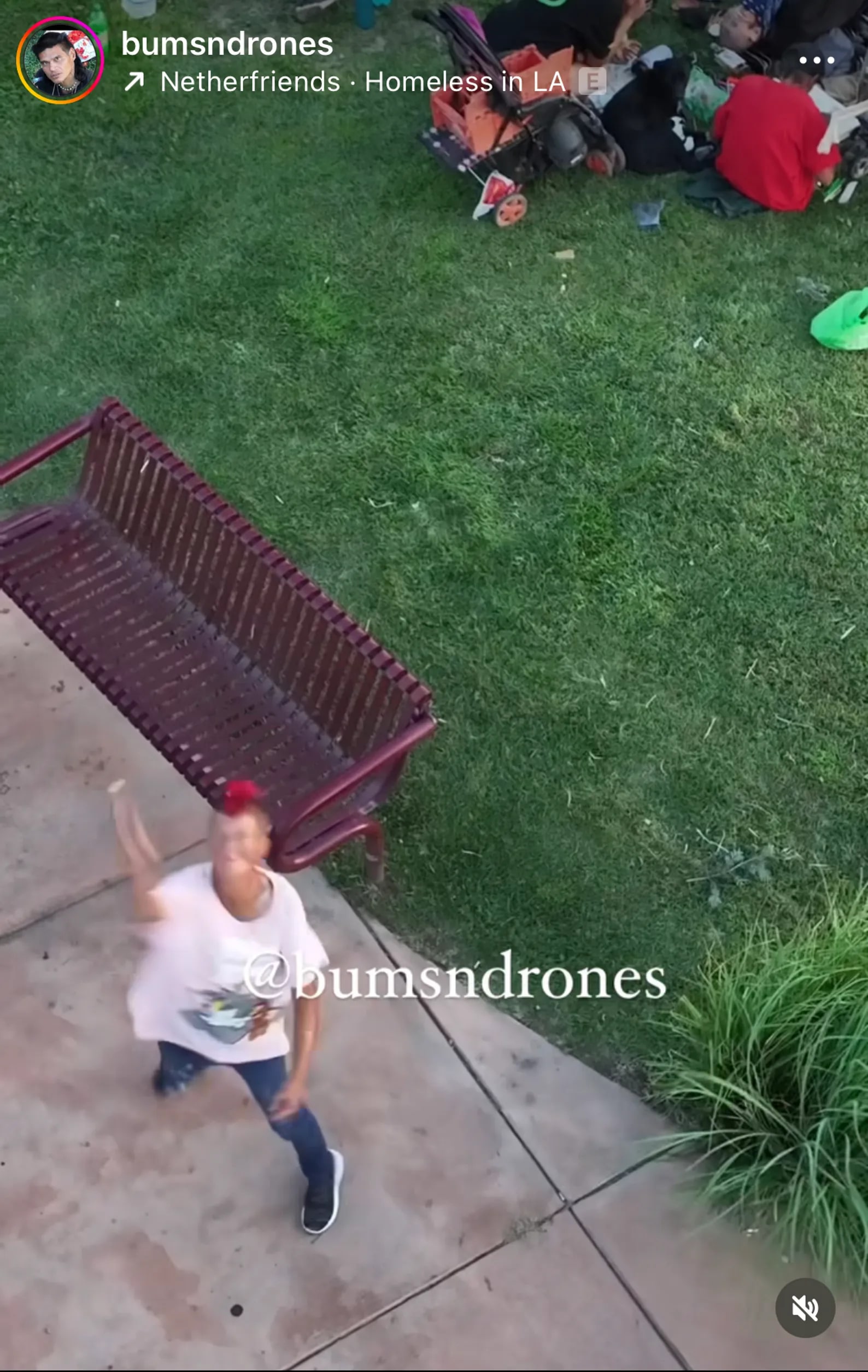 A person with a red-tinted hairstyle waves energetically toward the camera on a pavement near a maroon bench. Nearby, an area of grass has belongings and a person resting. The words "@bumsndrones" are visible on the image.