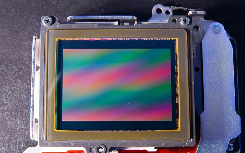 Close-up of a camera sensor displaying a colorful pattern with bands of green, pink, and blue. The sensor is mounted on a metallic frame, positioned against a dark background, showcasing intricate electronic components.