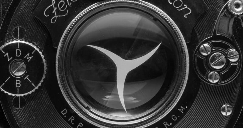 Close-up of an old camera lens with vintage markings. The black and white image highlights intricate details, including a prominent central icon resembling a metallic triangle. Surrounding the glass are mechanical dials and lettering, enhancing its classic look.