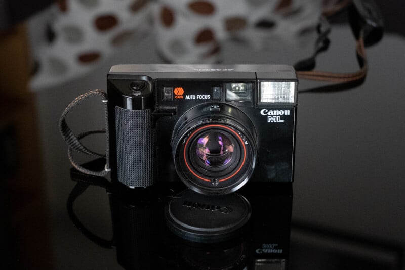 A vintage Canon AF35ML camera with a strap and lens cap on a reflective black surface. The camera features an auto-focus function and has a prominent flash on the upper right side.