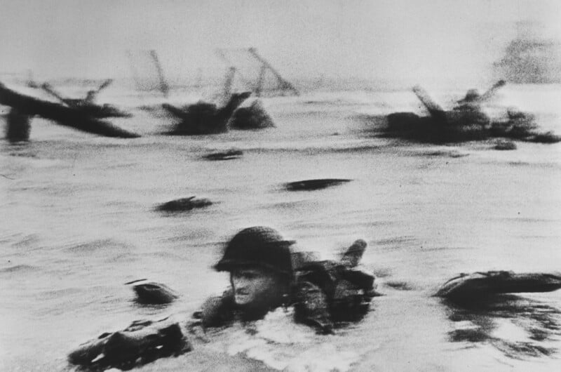 A soldier in uniform swims through choppy waters amidst WWII beach landings, with obstacles and structures visible in the background. The image is in black and white, conveying a sense of urgency and historical significance.