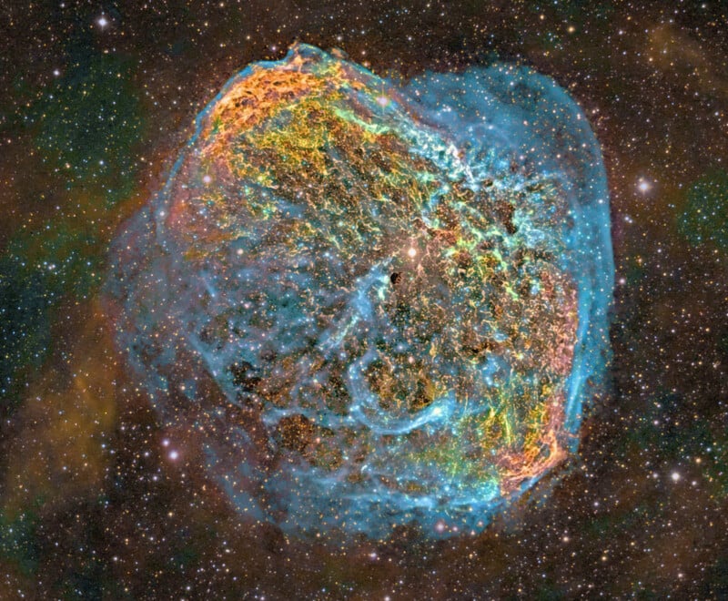 A vibrant, colorful nebula in space, displaying intricate patterns of blue, orange, and green gas clouds against a backdrop of stars. The nebula has a rounded, shell-like appearance with a textured surface.