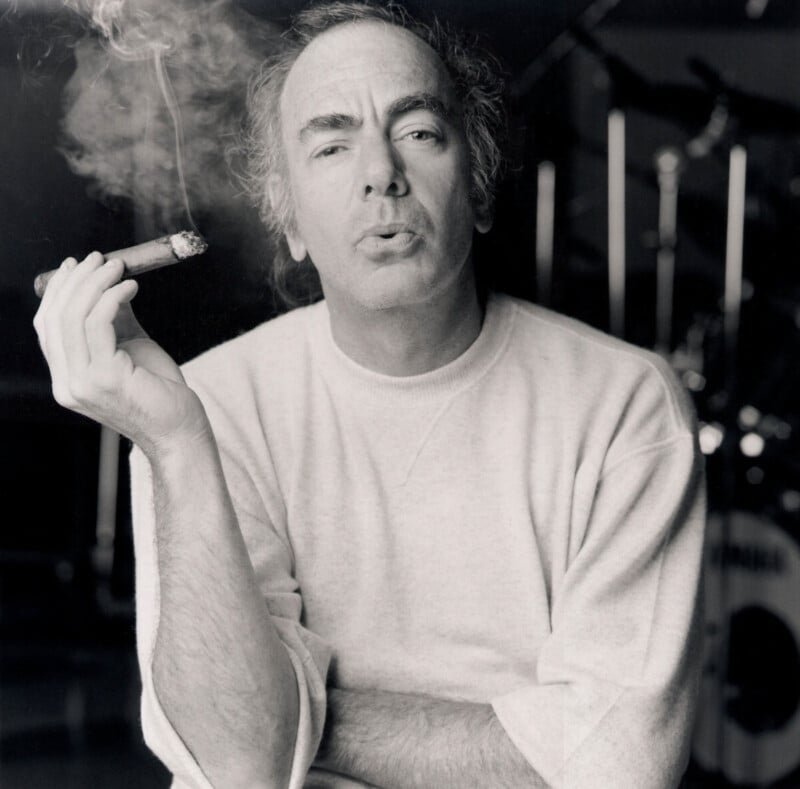 A black and white photo of a person with short hair wearing a light-colored sweater. They are holding a lit cigar, with smoke swirling around. The background features blurred musical instruments. The person appears relaxed and contemplative.