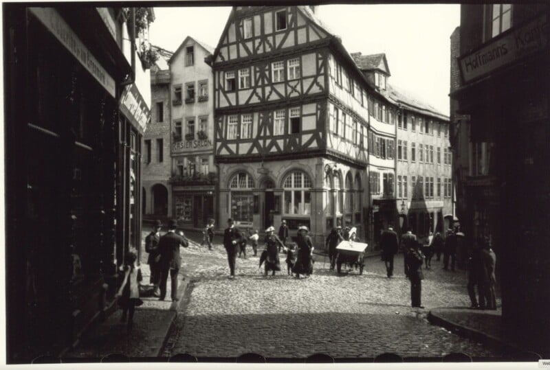 A black and white photo of a cobblestone street in a historic town. People walk and interact in front of half-timbered buildings with steep gabled roofs. The scene captures an old-world European ambiance with shops lining the street.