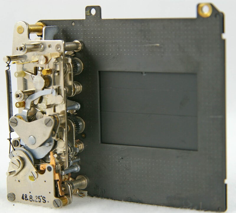 Close-up of a vintage mechanical part, possibly a camera shutter. It features intricate metal components and levers on a black rectangular frame. The assembly appears detailed with springs and gears visible.