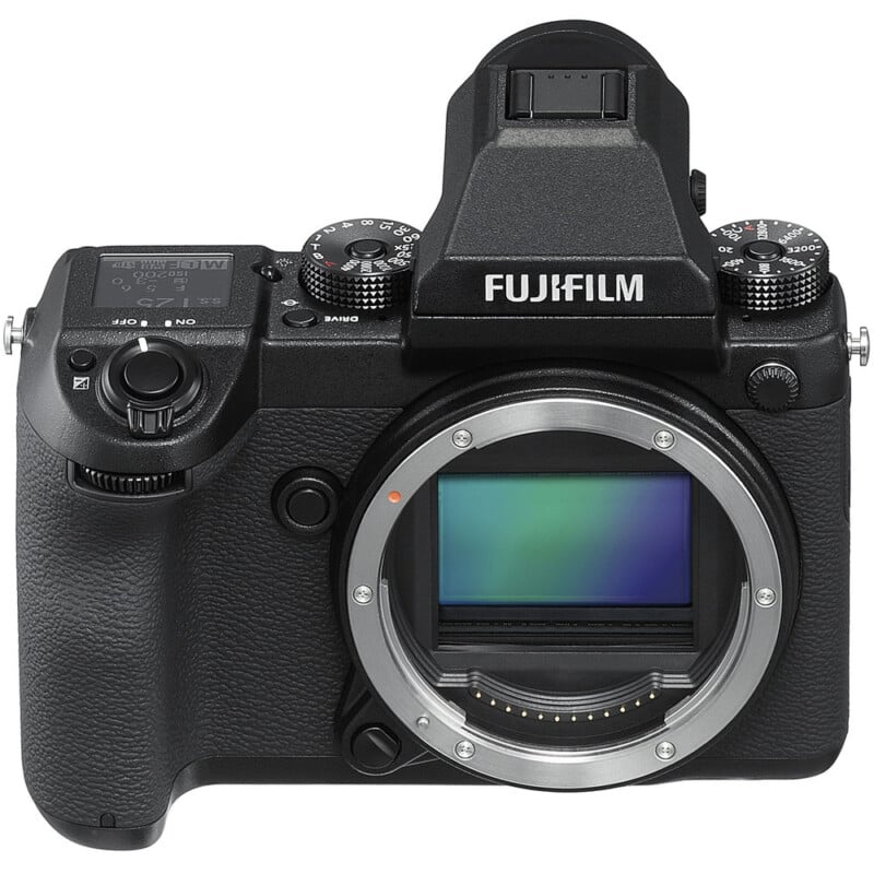 Front view of a Fujifilm digital mirrorless camera body without a lens attached, showing the sensor. The camera has several control dials on top and a textured grip on the right side.